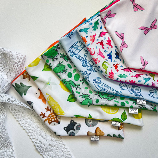 Six dog bandanas in woodland creatures, lemons, frogs, construction trucks, dinosaurs and pink bows overlap in a fan-like arrangement.