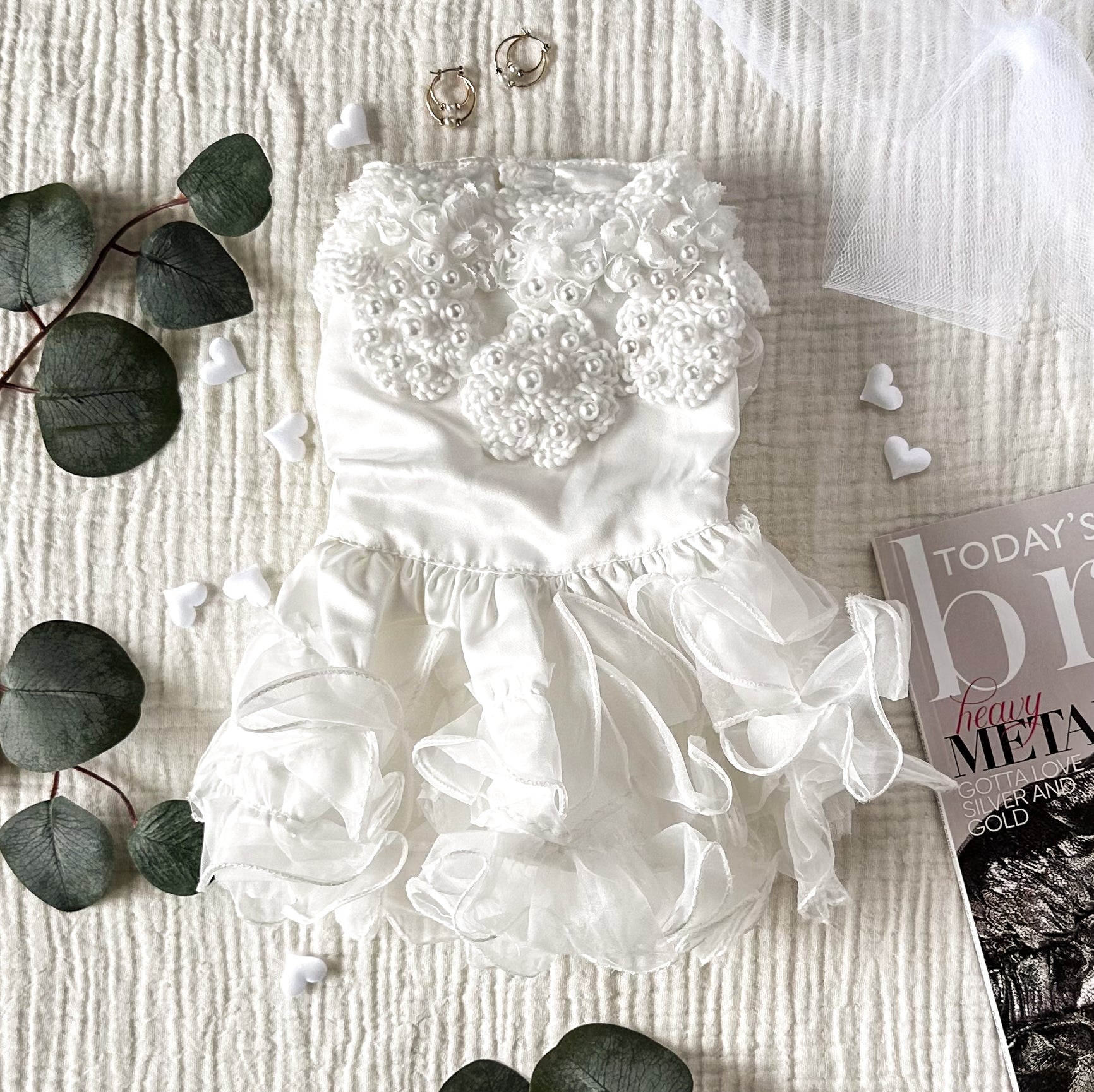 A white dog wedding dress with pearls and a tulle skirt on a textured white background, surrounded by gold earrings, white hearts, plant leaves and a magazine corner.