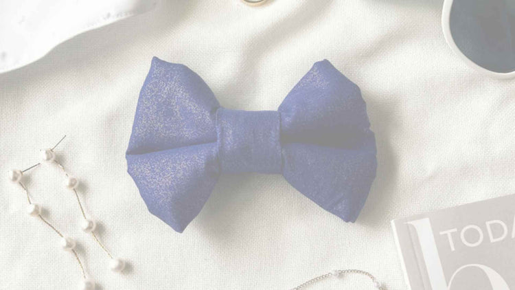 A blue sparkly dog wedding bow on an off-white background. The bow is surrounded by decorative items such as elegant gold jewelry, white sunglasses and the corner of a magazine.