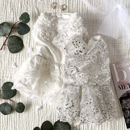 Two white dog wedding dresses, one with white pearls and a tulle skirt and the other with silver sequins. The dresses are laying on a white textured background and are surrounded by white hearts, gold earrings, plant leaves and a magazine corner.