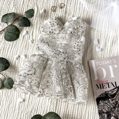 A white dog wedding dress with silver sequins on a textured white background, surrounded by gold earrings, white hearts, plant leaves and a magazine corner.