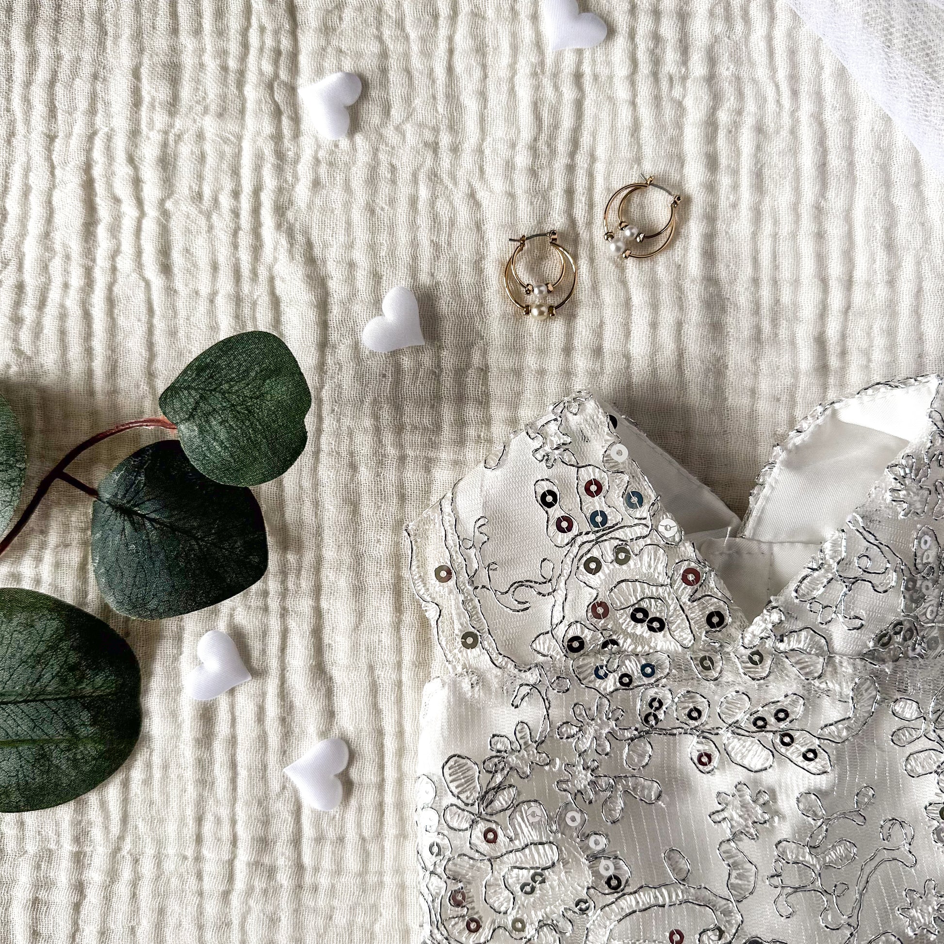 The top half of a white dog wedding dress with silver sequins on a white textured background, with gold earrings, plant leaves and white hearts.