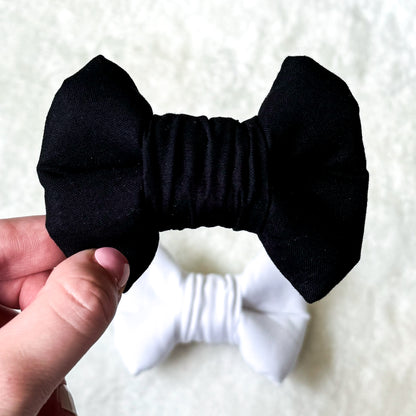 A hand holding a black formal dog bowtie in the foreground with a white formal dog bowtie in the background.