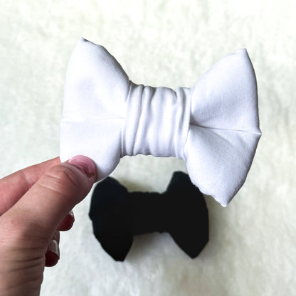 A hand holding a white formal dog bowtie in the foreground with a black formal dog bowtie in the background.